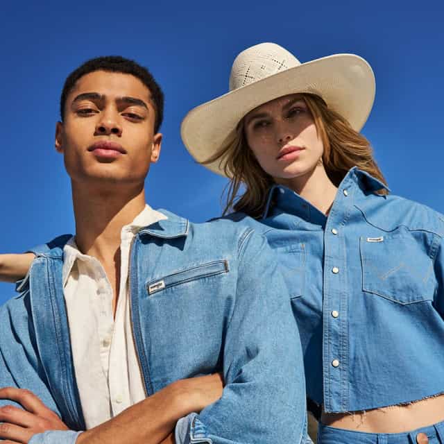 Image of couple wearing Wrangler. The man is on the left, wearing a white shirt and a light jean jacket. The woman is wearing a jean shirt, a cream cowboy hat, and jeans.